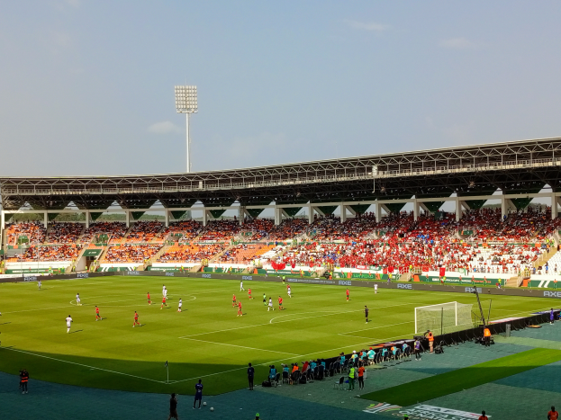 Ein Fußballspiel in einem großen Stadion mit Spielern auf dem Boden, einem Tor, einem Zaun, einer Fahne mit Text, Metallrahmen, einem Dach mit Deckenbeleuchtung und einem bewölkten Himmel.