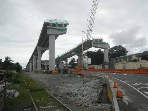 Baustelle mit einer Brücke im Hintergrund, Straße mit Absperrbaken, Eisenbahnschiene, verstreute Steine und Gras, Bäume und Gebäude auf beiden Seiten und ein bewölkter Himmel.