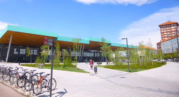 Eine Gruppe von Fahrrädern, die vor einem modernen Gebäude mit Glaswänden und Pfeilern in Rotterdam, Niederlande, geparkt sind, umgeben von Bäumen, Laternenmästen und Fahrzeugen auf der Straße, mit Menschen, die herumgehen und einem bewölktem Himmel im Hintergrund.