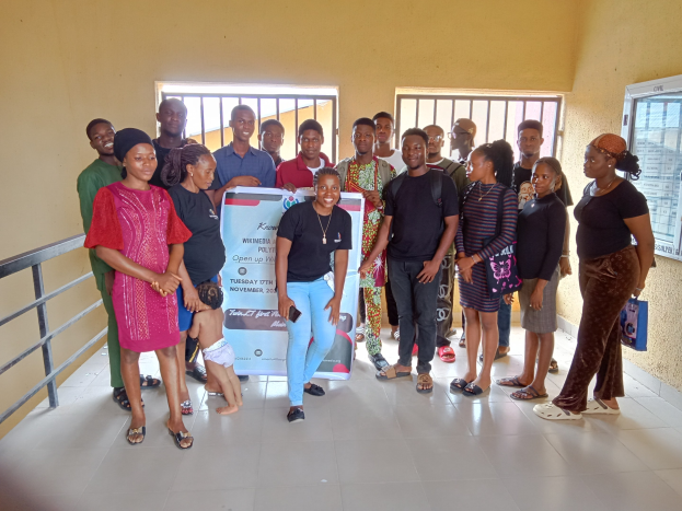 Eine Gruppe von Menschen, die zusammen in einem Raum stehen und eine Gemeindeinformationswand halten, mit einem Geländer auf der linken Seite, Fenstern und einer Wand im Hintergrund.