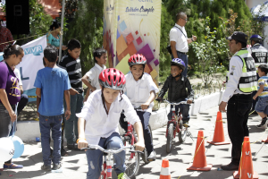Eine Gruppe von Kindern auf Fahrrädern, die eine Straße mit Verkehrskegeln entlangfahren, einige tragen Helme, andere stehen in der Nähe, mit einem Banner, Bäumen und Gebäuden im Hintergrund.