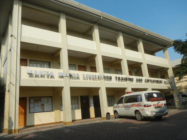 Lebensmittel-Trainings- und Arbeitsplatz-Zentrum-Santa-Maria-Gebäude-mit-Fenstern,-Türen-und-Text-umgeben-von-Bäumen-und-Fahrzeugen-auf-der-Straße-unter-einem-klaren-blauen-Himmel.