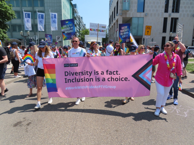 Eine bunte Gruppe von Menschen marschiert auf einer Stra├če, h├Ąlt ein Banner mit der Aufschrift "Vielfalt ist eine Tatsache, Inklusion ist eine Wahl" in den H├Ąnden, einige tragen K├Ąpse und Schutzbrillen, andere halten zus├Ątzliche Banner, vor einem Hintergrund von Geb├Ąuden, B├Ąumen und einem klaren blauen Himmel.
