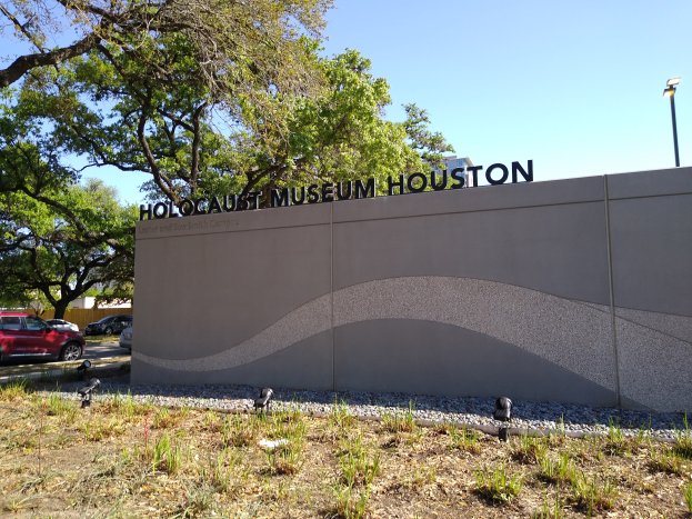 Das Holocaust-Museum in Houston, Texas, mit einer textbedeckten Wand, Steinen, Gras, Pflanzen, Bäumen auf der Straße, einem Straßenpfahl und einem bewölkten Himmel.