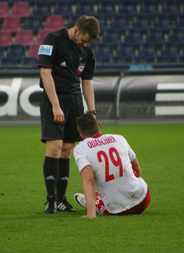 Ein Fussballspieler und Schiedsrichter sitzen auf dem Boden in einem Stadion, beide in Sportkleidung.