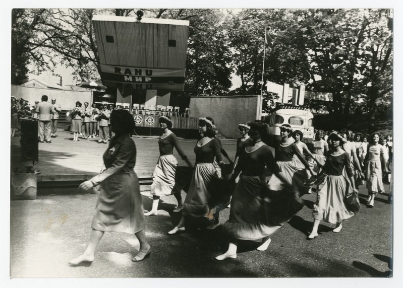 Eine Gruppe von Frauen tanzt auf der Straße, umgeben von einer Menge, einem Lautsprecher, einem Fahrzeug, einer Tafel mit Text und einer Fahnenstange.