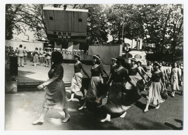 Eine Gruppe von Frauen tanzt auf der Straße, umgeben von einer Menge, einem Lautsprecher, einem Fahrzeug, einer Tafel mit Text und einer Fahnenstange.
