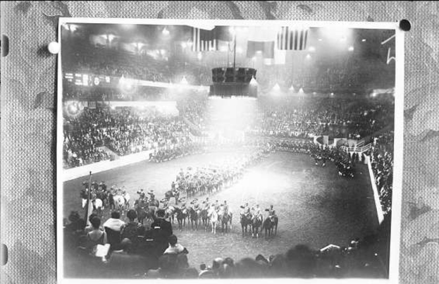 Ein Schwarz-Weiß-Foto einer großen Menschenmenge in einem Stadion, mit einigen auf Pferden und anderen stehend, eine zentrale Bühne mit Flaggen und Lichtern und Text unten.
