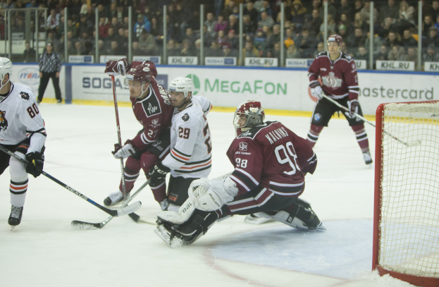 Gruppe von Menschen, die Hockey auf einem Eisstadion spielen, mit Torpfosten auf der rechten Seite, Helm und Stöcke tragend, Zuschauern in den Rängen mit Bannern im Hintergrund.