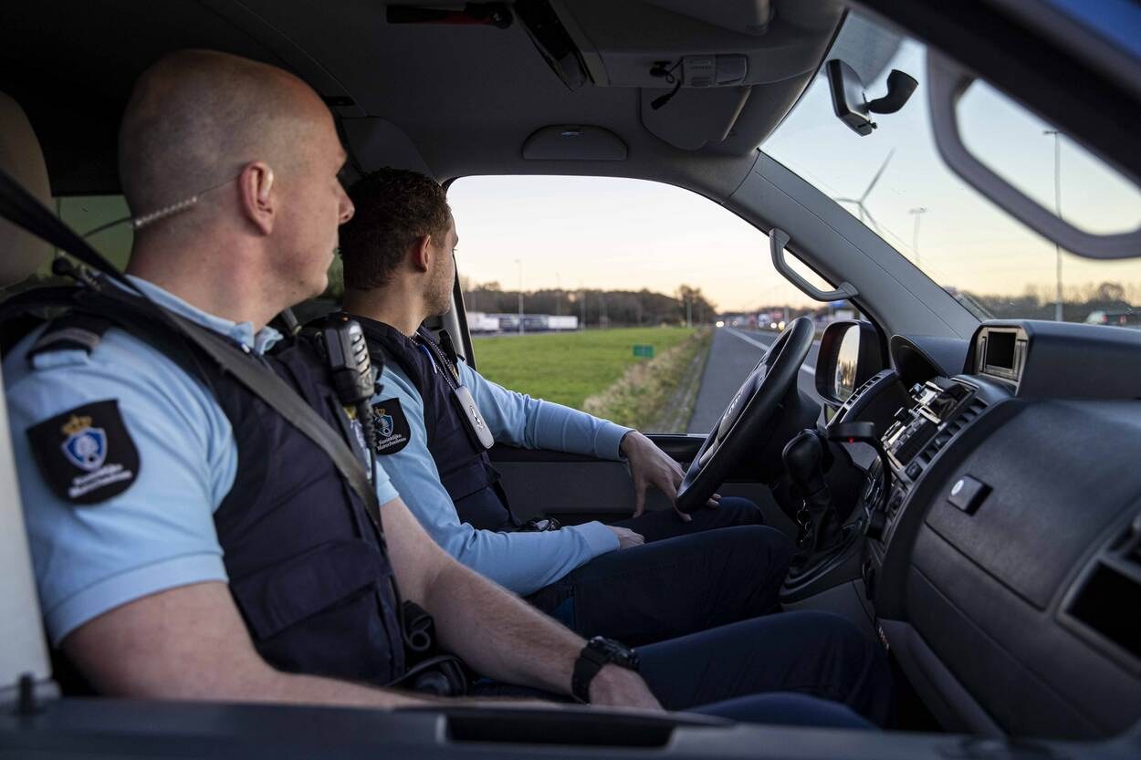 Zwei Polizeibeamte sitzen in einem Auto auf einer Straße, einer hält das Lenkrad, mit Gras, Bäumen und Pfählen im Hintergrund unter einem klaren blauen Himmel.