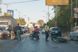 Eine Gruppe von Menschen steht um ein verunglücktes Motorrad auf der Straße herum, umgeben von mehreren Fahrzeugen, darunter ein Lastwagen, und einer Hintergrundlandschaft aus Bäumen, Pfählen, Lampen und Schildern unter dem Himmel.