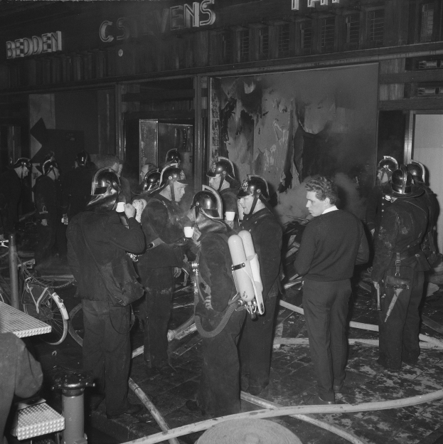 Schwarzes Bild von Feuerwehrleuten vor einem Gebäude, die Helme tragen und Sauerstoffflaschen tragen, mit einem Fahrrad und einem Tisch voller Gegenstände in der Nähe.