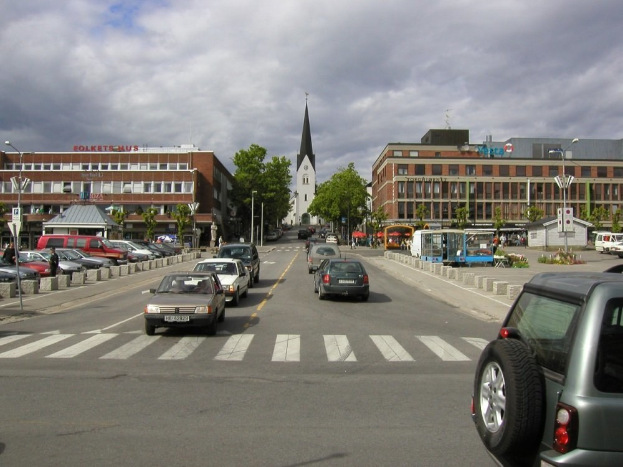 Stadtstraße mit parkenden Autos, Gebäuden, Bäumen, Laternen, einem bewölkten Himmel und einem sichtbaren Kfz-Kennzeichen.