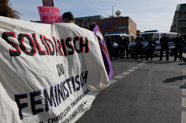 Eine Gruppe von Menschen marschiert mit einem Banner, auf dem 'Solidarität und Feminismus' steht, durch eine Straße, mit parkenden Fahrzeugen und Gebäuden im Hintergrund unter einem klaren blauen Himmel.