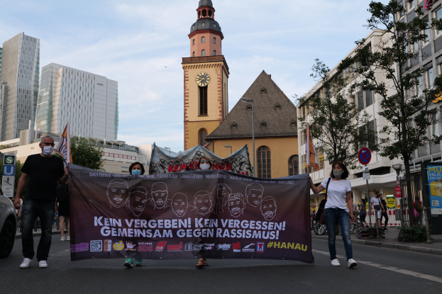 Eine Gruppe maskierter Menschen marschiert mit einem Banner die Straße entlang, mit einem geparkten Auto auf der linken Seite, Gebäuden, Bäumen, Schildern, Polen und einem Uhrenturm im Hintergrund unter einem klaren blauen Himmel.