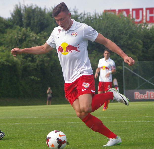 Ein professioneller Fussballspieler kickt einen Ball auf einem grünen Feld mit "RB Leipzig" im Hintergrund unter einem klaren blauen Himmel.