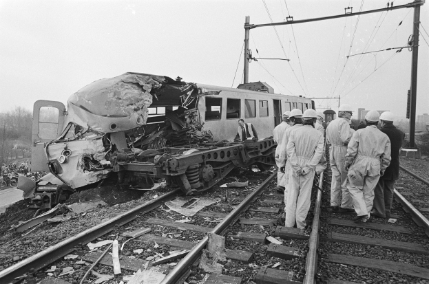 Schwarzes und weißes Bild einer Zugkollision auf den Schienen mit Einsatzkräften in Helmen in der Nähe, umgeben von Strommasten, Bäumen, Gebäuden und einem klaren Himmel.