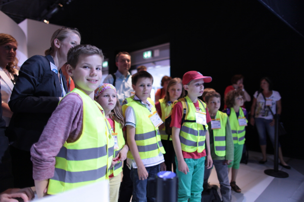 Eine Gruppe von Kindern mit Sicherheitswesten und Namensschildern, wobei ein Junge links lächelt, auf einem Boden mit einem Pfahl rechts steht und einige Menschen im Hintergrund zu sehen sind.