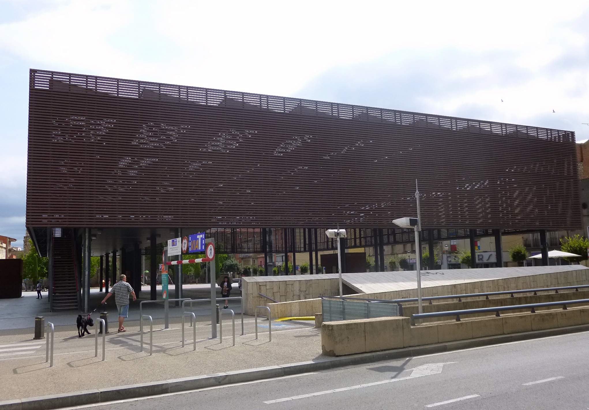 Großes modernes Gebäude mit zahlreichen Fenstern, das als neuer Eingang des Stadtzentrums dient, umgeben von Straßeninfrastruktur, Fahrzeugen, Fußgängern, Bäumen und einem bewölkten Himmel.