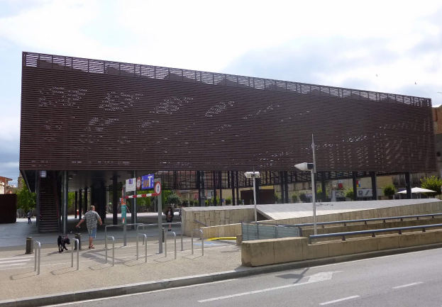 Großes modernes Gebäude mit zahlreichen Fenstern, das als neuer Eingang des Stadtzentrums dient, umgeben von Straßeninfrastruktur, Fahrzeugen, Fußgängern, Bäumen und einem bewölkten Himmel.