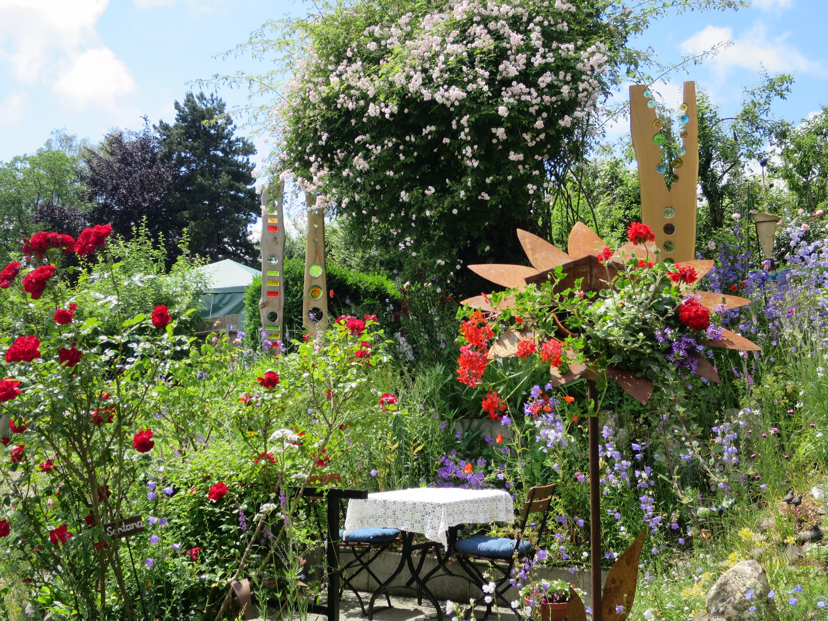 Ein Garten auf dem Chelsea Flower Show mit bunten Blumen und Pflanzen, einem Tisch und Stühlen in der Mitte mit einer Decke darauf, umgeben von Bäumen unter einem bewölkten Himmel.