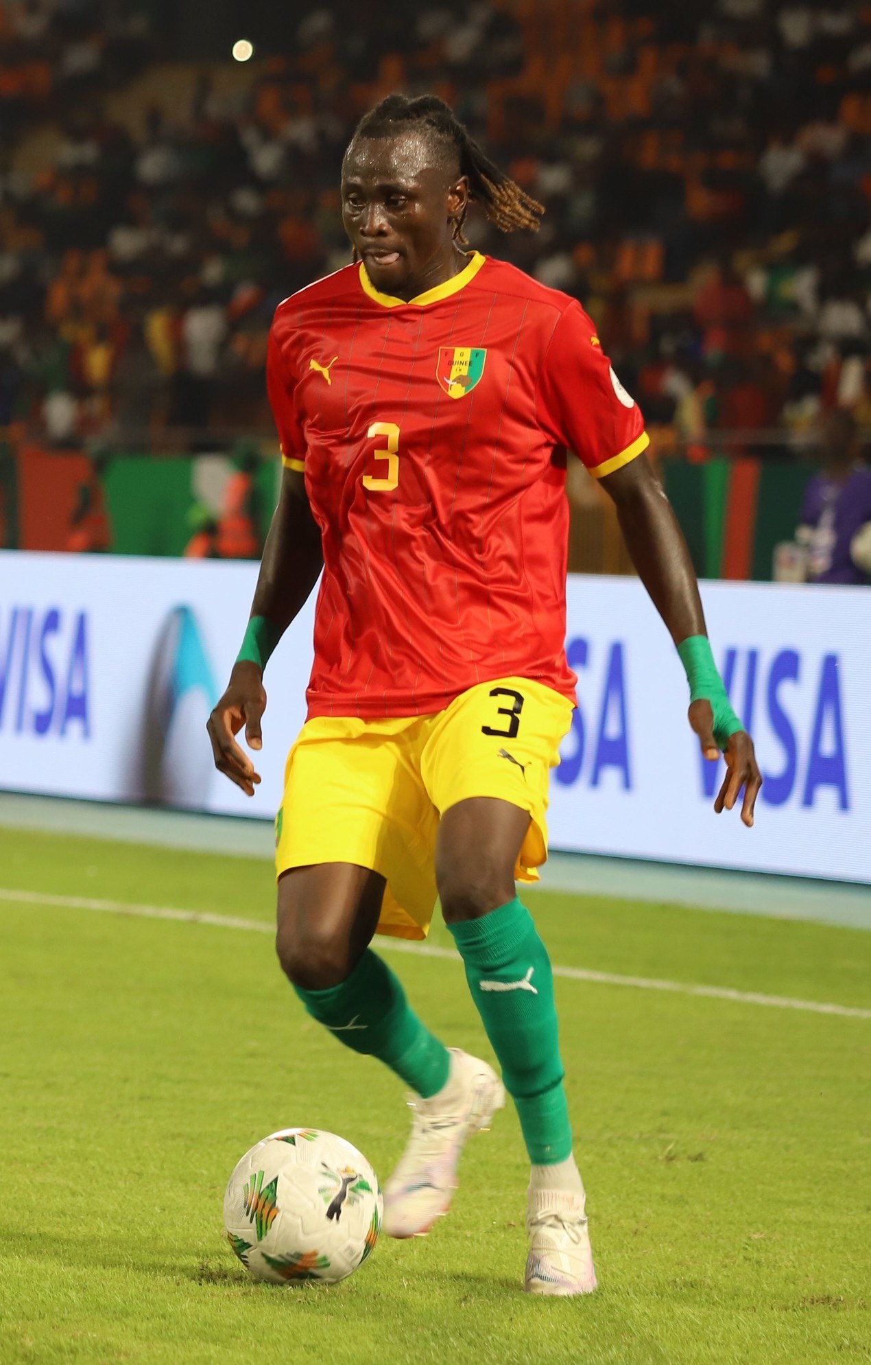 Ein Fussballspieler in rot-gelber Uniform, der einen Ball auf einem Feld mit Zuschauern in den Rängen und Werbetafeln im Hintergrund kickt.