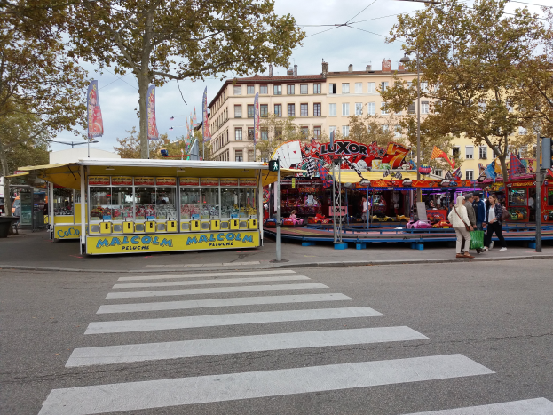 Eine belebte Stra√e mit Zebrastreifen, einer Karussellfahrt, Passanten, Ständen, Bäumen, Gebäuden, Bannern, Laternenmästen und einer Ampel unter einem bewölktem Himmel.