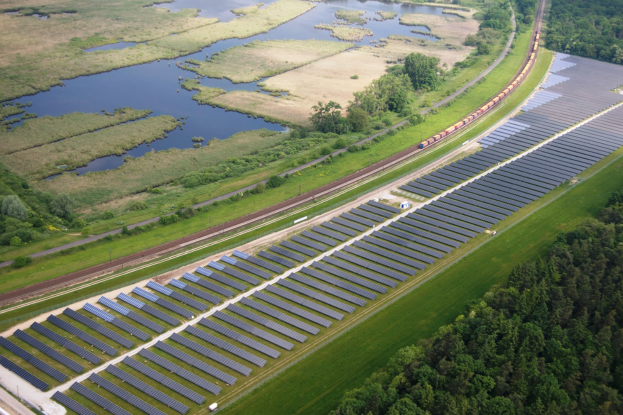 Luftaufnahme eines Solarparks mit Panelen in einem Feld, umgeben von Bäumen, Gras, Pflanzen und Wasser, mit einem Zug auf einer nahen Eisenbahnstrecke.