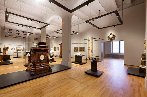 Ein geräumiger Raum in der National Gallery of Art, Washington, DC, mit verschiedenen Kunstwerken wie Skulpturen, Glasboxen, Säulen, an der Wand montierten Rahmen, einer Uhr, einem Fenster und Deckenleuchten.