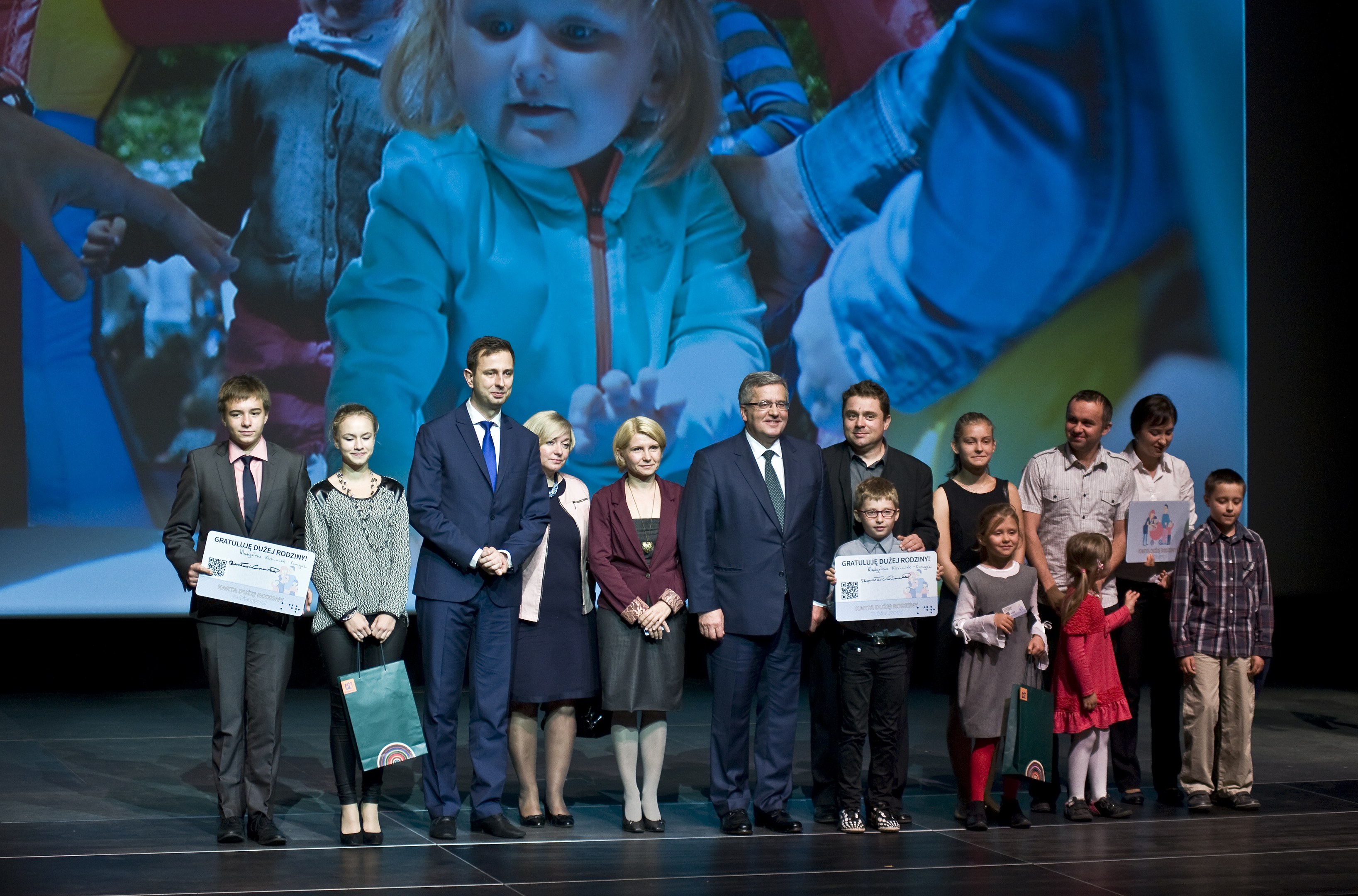 Gruppe von Menschen auf einer Bühne mit Taschen und Urkunden, im Hintergrund ein Bildschirm mit einem Bild eines Kindes und anderen.