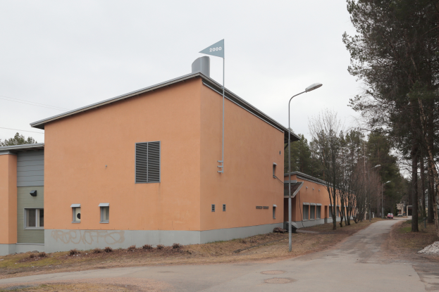 Ein orangefarbenes Gebäude mit einer Fahne darauf, umgeben von Straßenlaternen, Autos, Bäumen und einem klaren blauen Himmel.