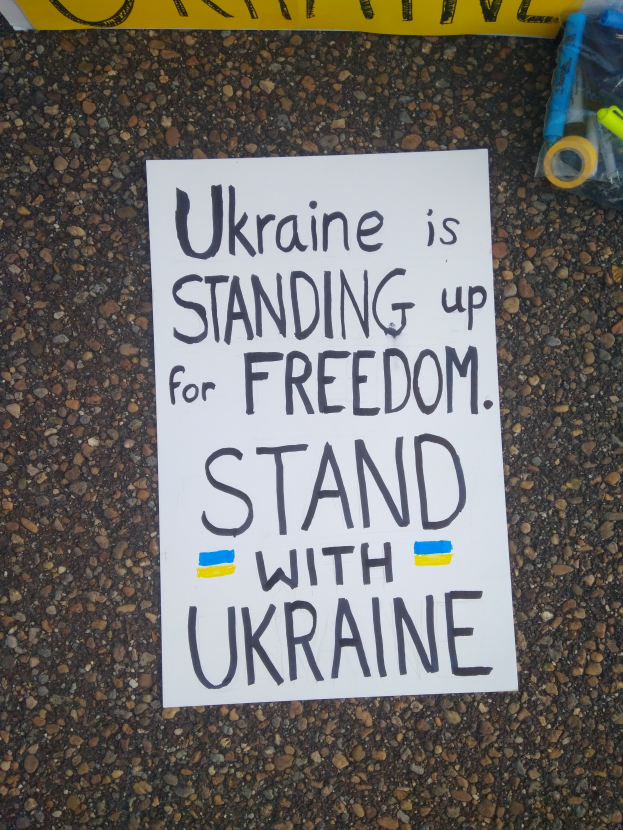 Ein weißer Ordner mit der Aufschrift "Ukraine steht für Freiheit, steht mit Ukraine" liegt vor einem gelben Banner auf dem Boden, rechts daneben eine Plastikabdeckung mit verschiedenen Gegenständen.
