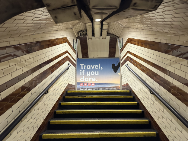 Eine Treppe in einer U-Bahn-Station mit einem "Reise, wenn du wagst"-Schild oben, flankiert von Geländern, mit Stationwänden im Hintergrund.