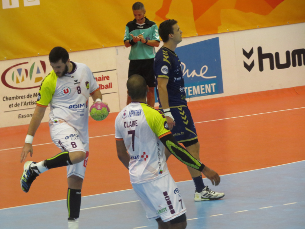 Eine Gruppe von Männern, die Handball auf einem Platz spielen, wobei ein Spieler den Ball hält, in einer professionellen Einrichtung mit Bannern im Hintergrund.