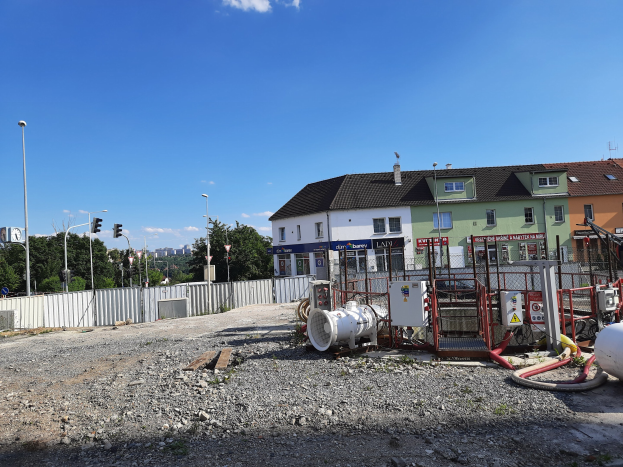 Baustelle mit freiliegenden Rohren und Geräten, umgeben von Gebäuden, Laternen, Ampeln, einem Zaun, Bäumen und einem bewölkten Himmel.