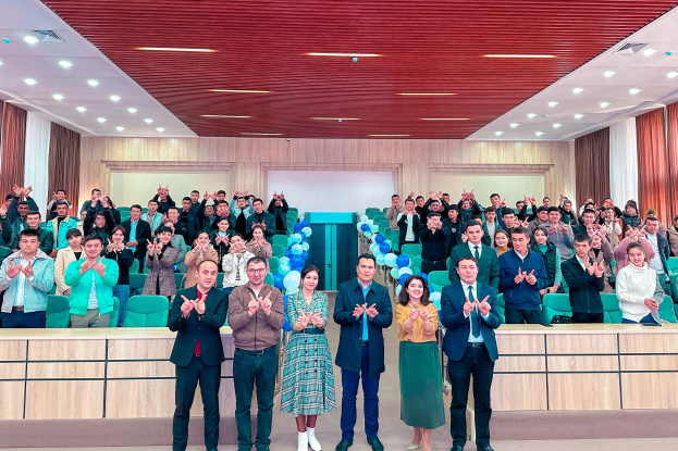 Eine Gruppe von Menschen steht auf einer Bühne vor einem Publikum, wobei einige Zuschauer klatschen, während der Eröffnungsfeier einer neuen Schule, mit Stühlen, Tischen, Luftballons, Vorhängen und Deckenleuchten im Hintergrund.