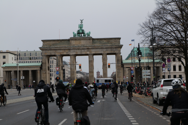Eine Gruppe von Menschen, die auf Fahrr√°dern die Stra√e vor dem Brandenburger Tor in Berlin, Deutschland, entlangfahren, mit B√§umen, Laternen, Verkehrszeichen, Schildern und Geb√§uden mit Fenstern im Hintergrund und einem sichtbaren Himmel.