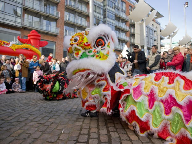 Ein lebendiges chinesisches Neujahrsfest in Amsterdam mit einem Löwen tanzen im Vordergrund und einer Menge Menschen, einige halten Kameras, die sich darum versammeln, vor einem Hintergrund aus Gebäuden, Laternenmasten und einem klaren blauen Himmel.