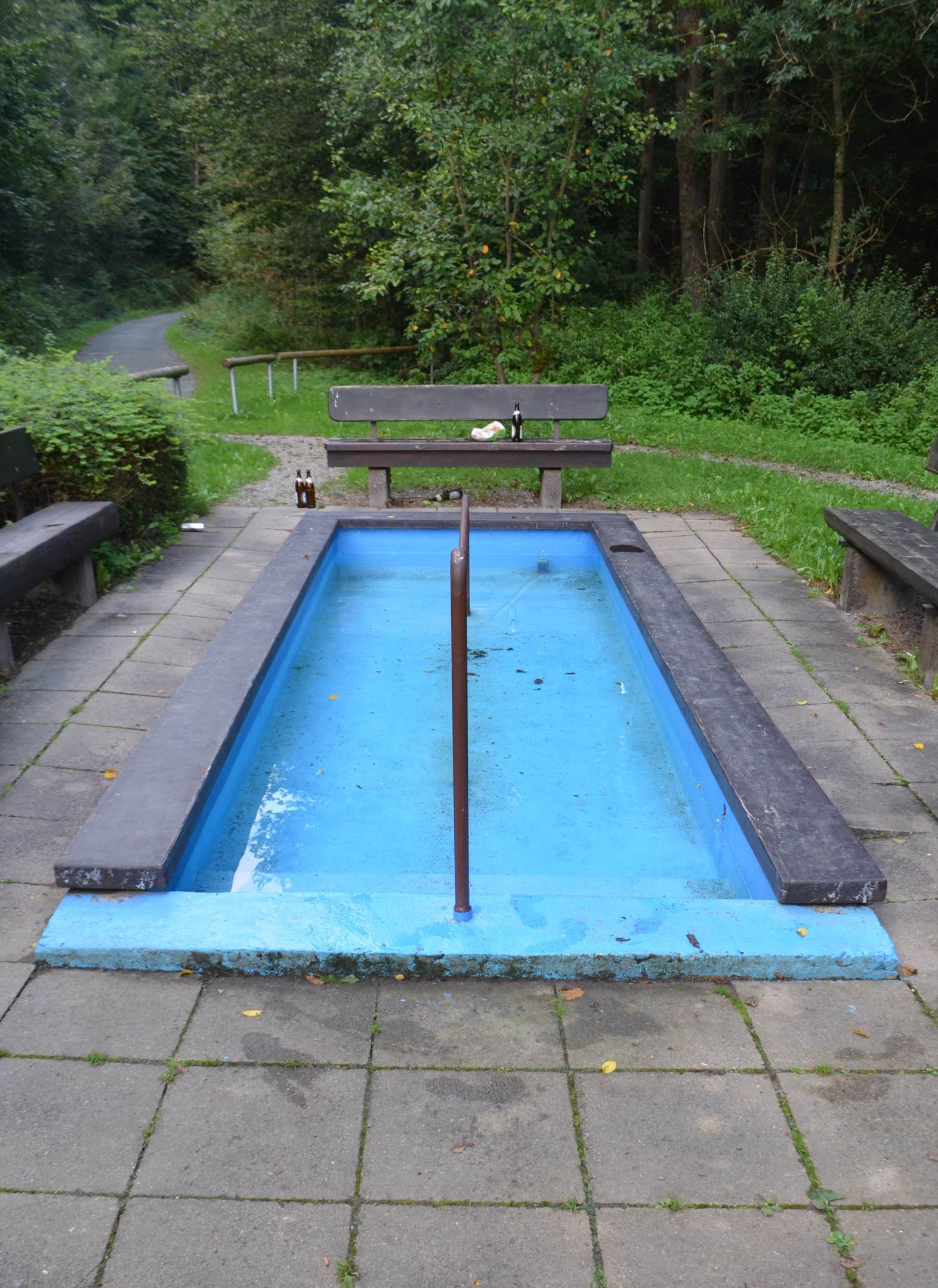 Kleiner Parkpool mit Wasser, zentralem Pfahl, umgeben von Bänken, Gras, Bäumen und einer Straße.