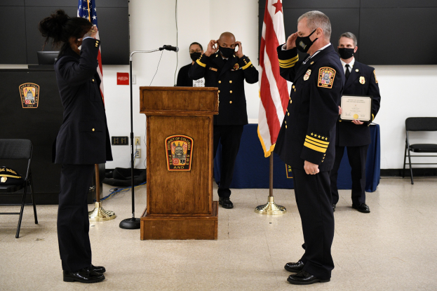 Gruppe uniformierter Personen in Masken vor einem Rednerpult während einer Vereidigung eines neuen Feuerwehrchefs, der eine Urkunde hält, umgeben von Flaggen, Stühlen, Tischen und einer Wand.