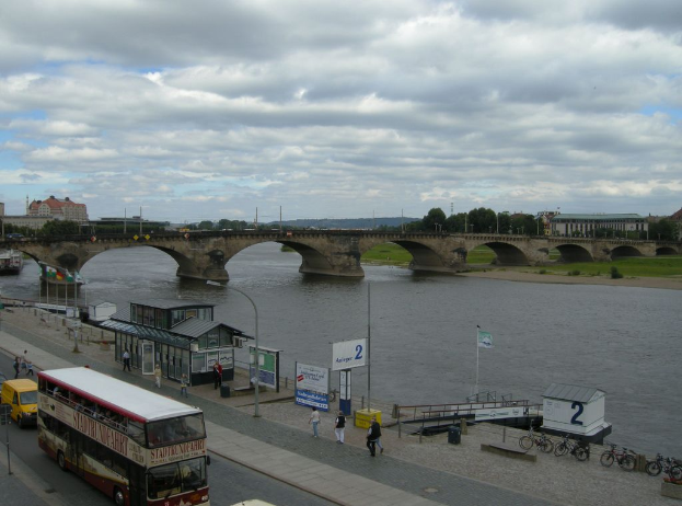 Eine belebte Straßenszene mit Fahrzeugen, Fußgängern, Fahrrädern, Gebäuden, einer Brücke und einem bewölkten Himmel, mit einem Plakat, einer Straßenlaterne, Stromkabeln und Gras.