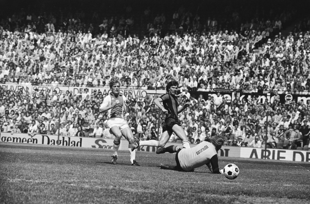 Ein Schwarz-Weiß-Bild einer Gruppe von Männern, die auf einem Feld Fußball spielen, mit Zuschauern im Hintergrund und einer Fahne mit der Aufschrift "Bundesliga 1978-79 Ajax 1-1 Hannover 96."