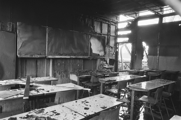 Schwarz-weiß-Foto eines Klassenzimmers mit verkohlten Wänden, zerbrochenen Fenstern und leeren Tischen und Stühlen.