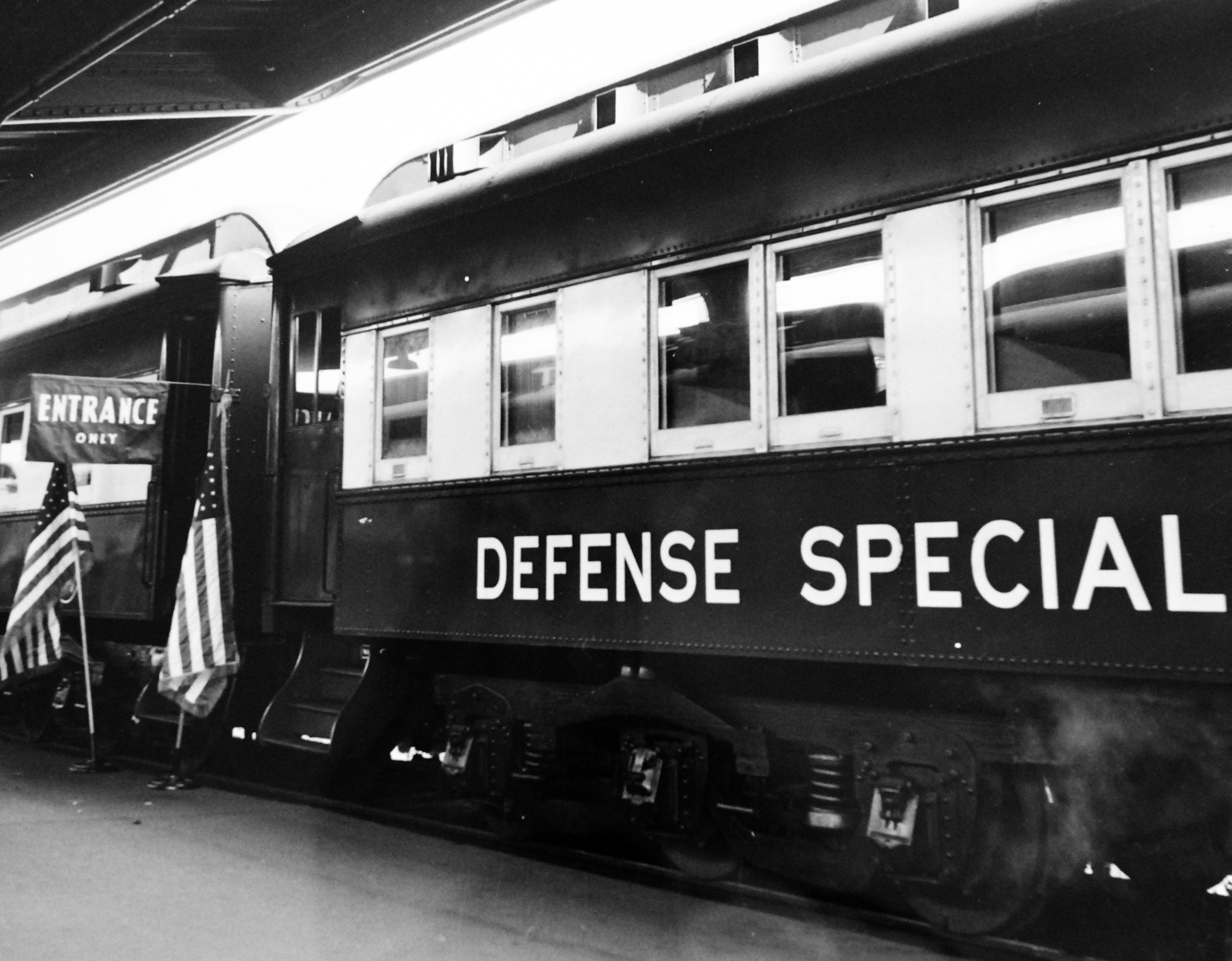 Schwarz-weiß-Foto eines Zuges mit der Aufschrift "Verteidigungsspezial" auf den Eisenbahnschienen mit zwei Fahnen links und einer Decke darüber.