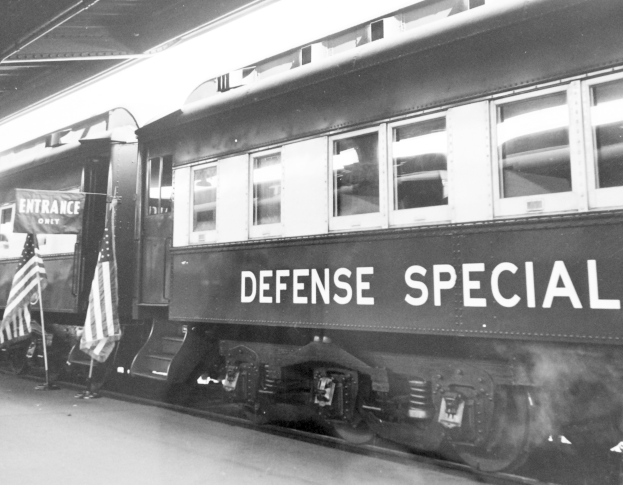 Schwarz-weiß-Foto eines Zuges mit der Aufschrift "Verteidigungsspezial" auf den Eisenbahnschienen mit zwei Fahnen links und einer Decke darüber.