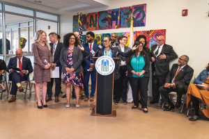 Gruppe von Menschen vor einem Rednerpult mit Mikrofon und einer Tafel mit Text, einige sitzen auf Stühlen und andere stehen, mit Flaggen, einer Tafel, einem Feuerlöscher und Deckenleuchten an der Rückwand.