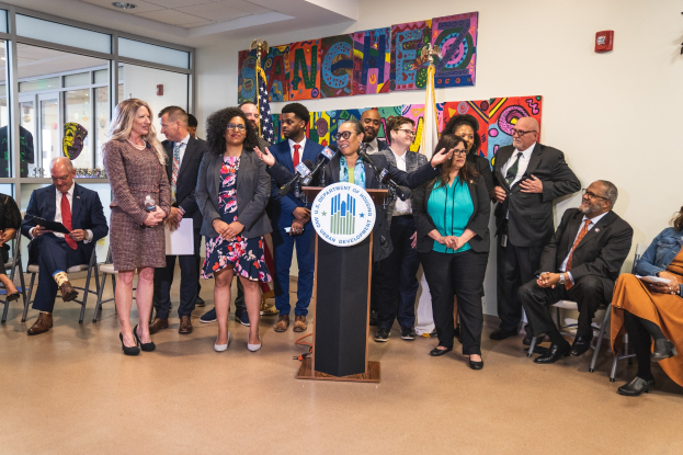 Gruppe von Menschen vor einem Rednerpult mit Mikrofon und einer Tafel mit Text, einige sitzen auf Stühlen und andere stehen, mit Flaggen, einer Tafel, einem Feuerlöscher und Deckenleuchten an der Rückwand.