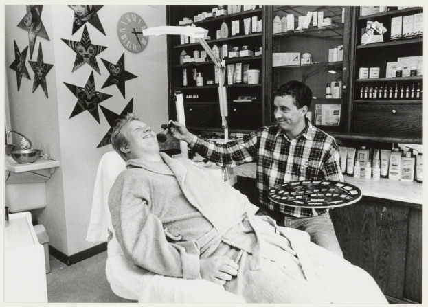 Ein Mann sitzt lächelnd in einem Zahnarztstuhl, während ein anderer Mann neben ihm steht und ein Tablett hält, mit einem Waschbecken, Sternen, einer Uhr und Regalen voller Flaschen im Hintergrund.
