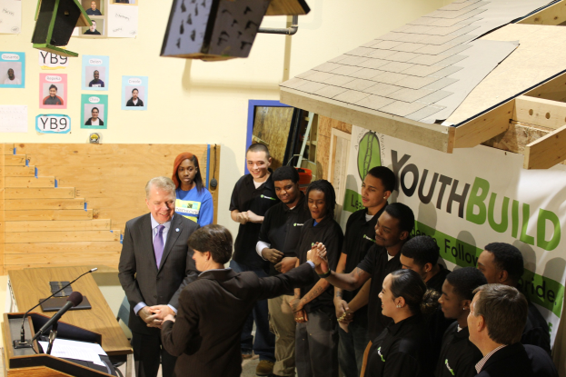 Gruppe von Menschen, die sich die Hand geben, um einen Podium mit Mikrofonen und Papieren auf einem Tisch, vor einer Menge, mit Plakaten an der Wand und Gegenständen auf dem Dach und der rechten Seite.