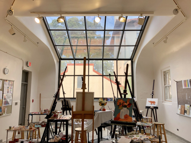 Ein unordentliches Kunstatelier mit Gemälden auf Staffeleien, Tischen und Hockern, mit einer Glaswand, die auf Bäume, Gebäude und den Himmel blickt, Teil des Art Institute of Chicago's neuen Studio.